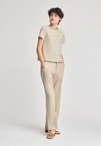 Beige kortärmad t-shirt med texten "Polo Club" och beige skräddarsydda byxor. Modellen står med händerna i fickorna och bär bruna sandaler. Slät tygtextur.