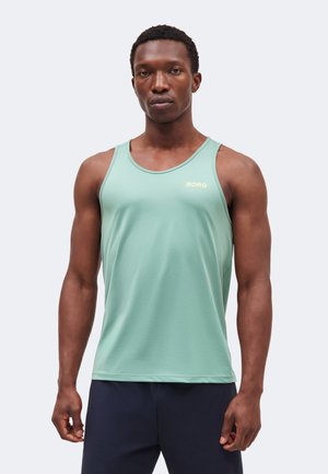 Muž s krátkymi kučeravými vlasmi, ktorý má na sebe svetlozelené športové tielko bez rukávov a tmavé šortky, čelí pred jednoliatym pozadím.