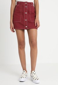 Jupe en denim rouge avec des boutons sur le devant et des détails de couture blancs, présentant une silhouette ajustée et deux poches avant. Associée à des chaussures à pois.