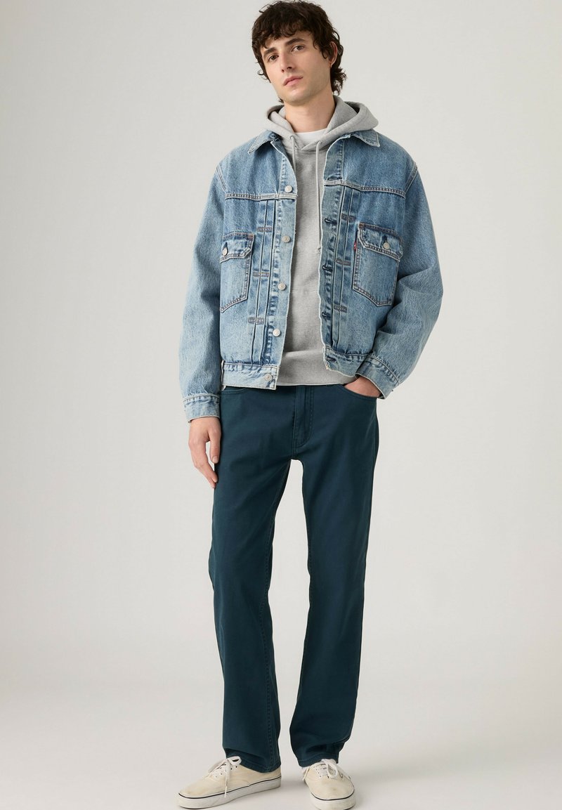 Junger Mann, der eine hellblaue Jeansjacke über einem grauen Hoodie, marineblaue Hose und weiße Turnschuhe trägt und mit einer Hand in der Tasche steht.