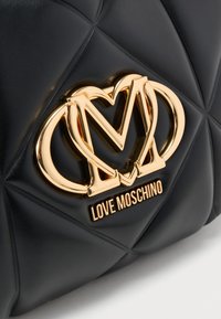 Μαύρη τσάντα από ψεύτικο δέρμα με σχέδιο καπιτονέ, με χρυσή διακοσμητική πλεγμένη λογότυπο και όνομα μάρκας, με ομαλή υφή και εμφανή διακοσμητικά στοιχεία.