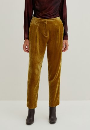 Femme portant un pantalon en velours jaune moutarde associé à des bottines marron foncé et un haut à manches longues bordeaux foncé.