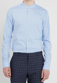 Light blue button-up shirt with a mandarin collar, made of smooth fabric, paired with dark plaid trousers.
