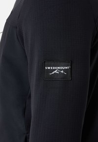 Zwarte textieljack met een gestructureerd oppervlak, voorzien van een rechthoekige stoffen patch met het "SWEDEMOUNT"-logo in het wit.