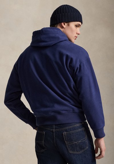 Muž so sivomodrou mikinou s kapucňou, tmavými džínsami a čiernou pletenou čiapkou, sediaci bokom na jednotnom béžovom pozadí.