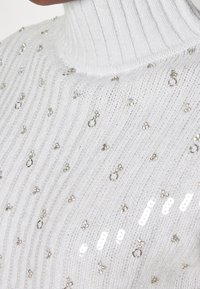 Pull à col roulé en tricot blanc avec des côtes verticales, orné de sequins argentés et d'embellissements circulaires épars.
