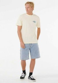 Lichtgeel T-shirt met korte mouwen en een blauw logo op de borst, gecombineerd met blauwe spijkershorts en zwarte sneakers met witte veters.