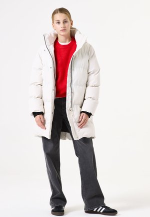 Weißer Daunenmantel mit hohem Kragen, Reißverschluss und übergroßer Passform. Ergänzt durch einen roten strukturierten Pullover und dunkle, weit geschnittene Jeans.