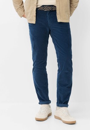 Uomo che indossa pantaloni di velluto a coste blu, sneakers bianche, camicia beige e cintura intrecciata, in piedi con le mani nelle tasche su sfondo bianco.