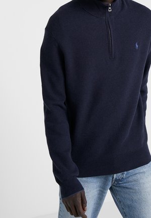 Man wearing navy blue knit quarter-zip sweater with small logo on chest and light blue jeans against plain background.