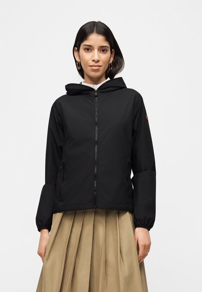 Jeune femme portant une veste à capuche noire zippée et une jupe plissée beige, debout devant un fond blanc uni.