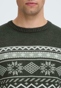 Pulover verde tricotat cu guler rotund cu nervuri, având un model geometric în crem și gri, incluzând forme de fulgi de zăpadă și diamante.