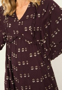 Lila Kleid mit V-Ausschnitt, fließenden Ärmeln und Rüschendetail. Hat einen metallischen Golddruck mit abstrakten Formen über den Stoff.