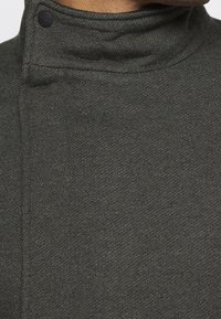 Tissu tricoté texturé vert foncé avec un col montant, doté d'un bouton noir pour la fermeture, présenté en vue rapprochée.