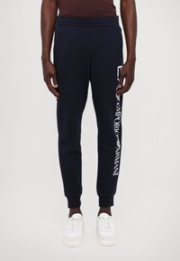 Tengerészkék jogger nadrág, rugalmas derékkal, a bal lábszáron fehér "Emporio Armani" logóval. Fehér sportcipővel párosítva.