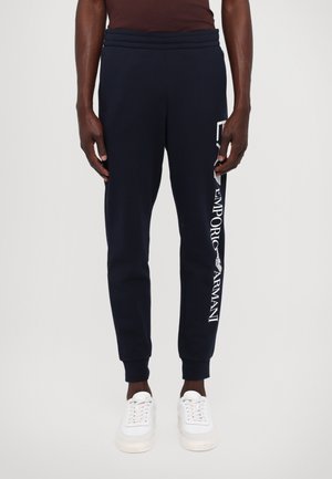 Joggers blu navy con vita elasticizzata, caratterizzati da un logo bianco "Emporio Armani" lungo la gamba sinistra. Abbinati a sneakers bianche.