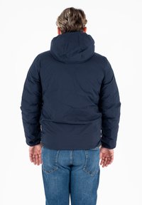 Giacca imbottita blu navy con cappuccio, caratterizzata da una texture liscia e un design imbottito. Indossata con jeans di denim, che mostrano una vestibilità casual vista da dietro.