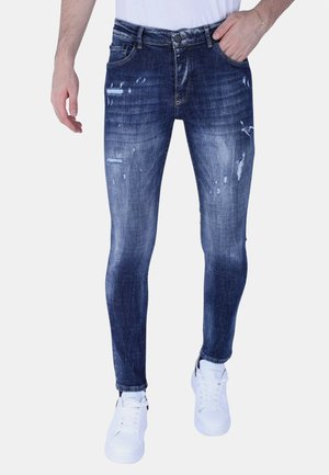 Man draagt slim-fit donkerblauwe versleten spijkerbroek en witte sneakers, staat met één hand in zak en andere ontspannen langs het lichaam.