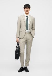 Άνδρας με μπεζ κοστούμι, λευκό πουκάμισο και πράσινη γραβάτα που κρατά μαύρη χαρτοφύλακα, στέκεται μπροστά σε απλό λευκό φόντο.