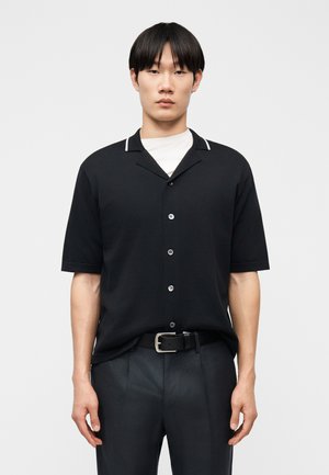 Muž oblečený v čiernej košeli s krátkym rukávom, zapínanej na gombíky, cez biele tričko a čierny opasok s tmavými nohavicami, stojaci pred jednoduchým svetlým pozadím.