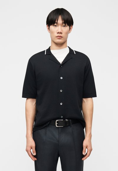 Homme portant une chemise noire à manches courtes boutonnée par-dessus un t-shirt blanc et une ceinture noire avec un pantalon foncé, debout devant un fond clair uni.