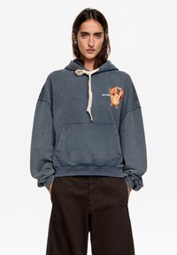 Niebieska, oversize'owa bluza z miękkiego materiału, z przednią kieszenią kangurzą, kapturem z regulowanym sznurkiem oraz kolorową grafiką psa na lewej piersi.