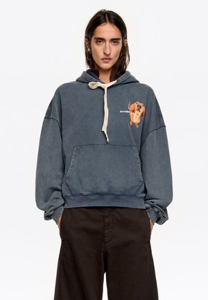 Niebieska, oversize'owa bluza z miękkiego materiału, z przednią kieszenią kangurzą, kapturem z regulowanym sznurkiem oraz kolorową grafiką psa na lewej piersi.