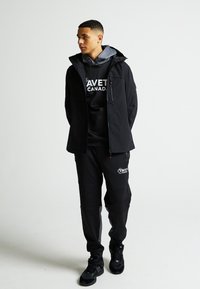 Černá voděodolná bunda přes šedou mikinu s logem, v kombinaci s černými tepláky a černými teniskami. Minimalistický design, praktické detaily.