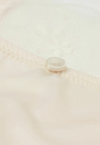 Lichtbeige stof met een gladde textuur, met een kleine, met stof bedekte knop dicht bij de gestikte rand. Achtergrond wit met een vaag patroon.