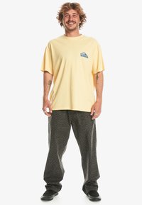 Camiseta de algodón amarilla con un pequeño logo azul en el pecho, combinada con pantalones relajados negros con un patrón y zapatos negros.