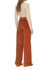 Weite Cordhose aus gestreiftem Wolle in verbranntem Orange, mit hoher Taille und strukturierter Oberfläche. Kombiniert mit einem langärmeligen gestreiften Oberteil.