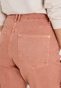Jean en denim rose clair à taille haute, avec des poches arrière et des détails de couture visibles. Le tissu a une texture lisse.