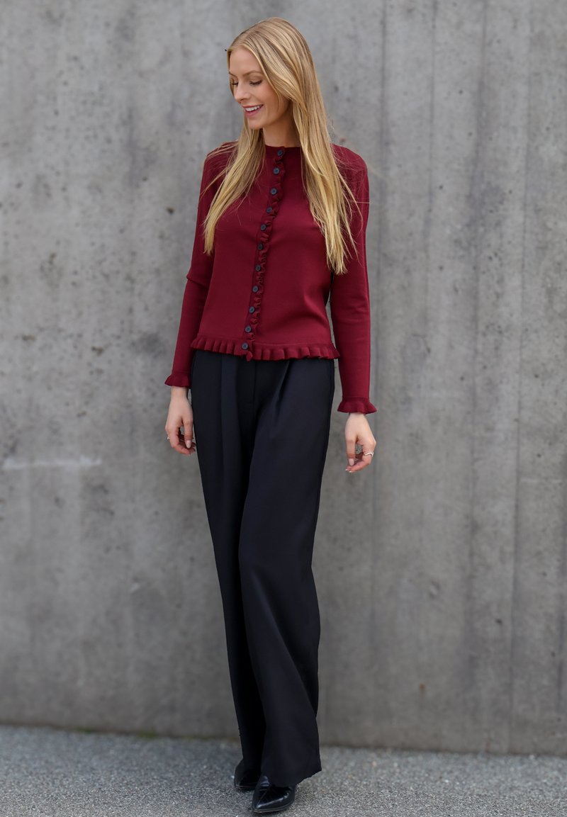 Femme aux longs cheveux blonds portant un cardigan bordeaux boutonné et un pantalon large noir, debout contre un mur en béton gris.