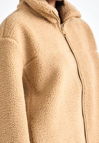 Close-up of beige zip-up fleece jacket with textured, fluffy fabric and high collar.