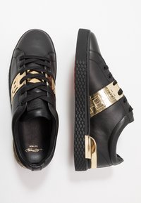 Zapatillas de cuero negro con acentos metálicos dorados en los laterales y el talón, cordones negros y suelas de goma texturizada negra sobre fondo blanco.