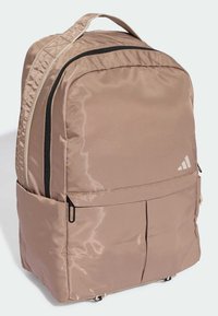 Tan spalvos kuprinė su lygiu, blizgiu audiniu. Yra juodi užtrauktukai, priekinė kišenė ir siuvinėta rankena viršuje. Baltas Adidas logotipas.
