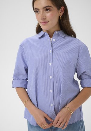 Jauna moteris dėvi mėlynai baltą vertikalių juostų marškinius su atlenktomis rankovėmis, auksinius lankelio formos auskarus ir kelis auksinius apyrankes.