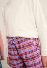 Kind dat een witte lange mouwen top en paarse geruite broek draagt, met een paarse strik bij de elastische tailleband, staande binnen.