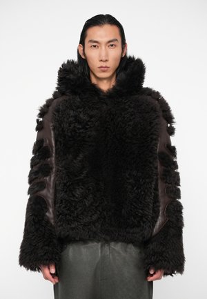 Rjava jakna iz ovčjega krzna z teksturirano, puhasto zunanjostjo in usnjenimi detajli; ima kapuco in izrezane detajle na rokavih.