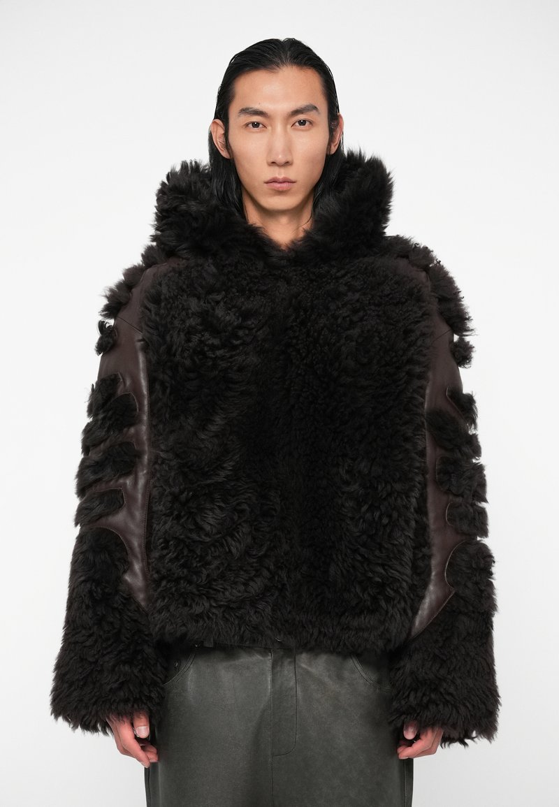 Brūns šerlinga mētelis ar teksturētu, pūkainu ārējo virsmu un ādas akcentiem; ar kapuci un izgriezumiem uz piedurknēm.