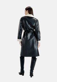 Cappotto di pelle nera con colletto e polsini in pelliccia sintetica, vita con cintura e maniche lunghe. Indossato sopra pantaloni neri aderenti e stivali con tacco.