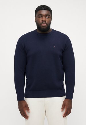 Granatowy sweter dzianinowy z okrągłym dekoltem, prążkowanymi mankietami i dołem, z małym logo na piersi. Noszony z kremowymi spodniami.