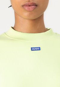 Svetlozelený rebrovaný pletený top s vysokým golierom a modrou visačkou s nápisom "HUGO" na hrudi. Hladká textúra, priliehavý strih.