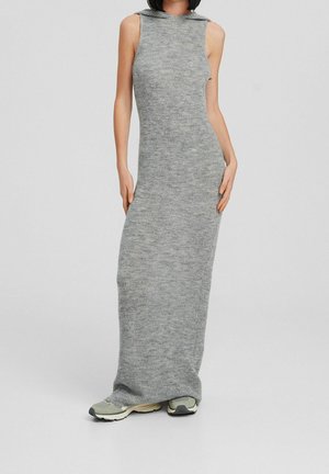 Femme portant une robe maxi en maille grise sans manches avec capuche, associée à des baskets gris clair, debout devant un fond clair uni.