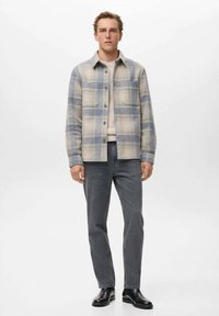 Karirana flanelska košulja u bež i svijetloplavoj boji, s raširenim ovratnikom, prednjim kopčanjem na dugmad i dvije džepove na prsima. Uparena s sivim trapericama.