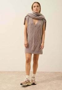 Sweter dzianinowy w jasnym mauve z głębokim dekoltem w kształcie litery V i wzorem w warkocze; zestawiony ze sportowymi snekerami i pasującym szalikiem luźno narzuconym na ramiona.