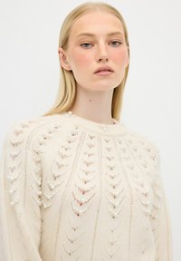Maglione lavorato a maglia in una tonalità crema, con un design in pizzo e dettagli in perle lungo le spalle e il collo. Tessuto morbido e scollatura rotonda.
