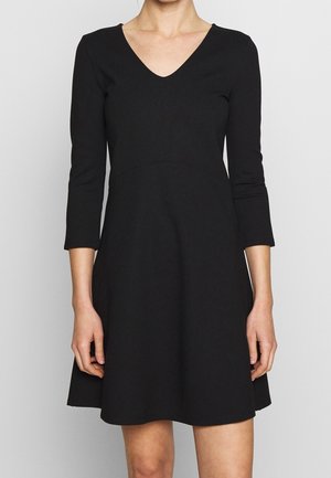 Mujer con un vestido negro hasta la rodilla, de manga tres cuartos y escote en V, posando frente a un fondo gris claro y liso.