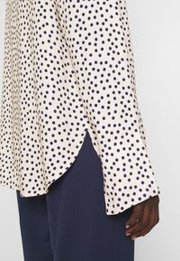 Blusa beige chiaro con pois blu scuro, maniche lunghe a polsino, orlo curvo, indossata con pantaloni blu scuro e mano visibile.