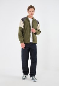 Giacca in pile verde oliva con accenti crema e grigio scuro, chiusura con zip e colletto alto. Abbinata a jeans blu scuro e sneakers.